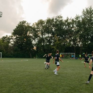 Women having an outdoor soccer game on a grassy field surrounded by trees.