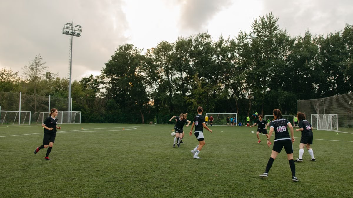 Women having an outdoor soccer game on a grassy field surrounded by trees.